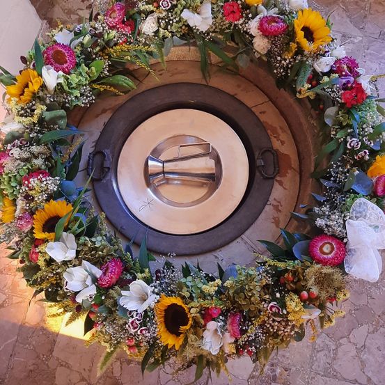 Eine kreisförmige Anordnung verschiedener Blumen umgibt ein metallisches Objekt, möglicherweise ein Feuerkorb, in der Mitte. Die Blumen sind in verschiedenen Farben, Formen und Größen.