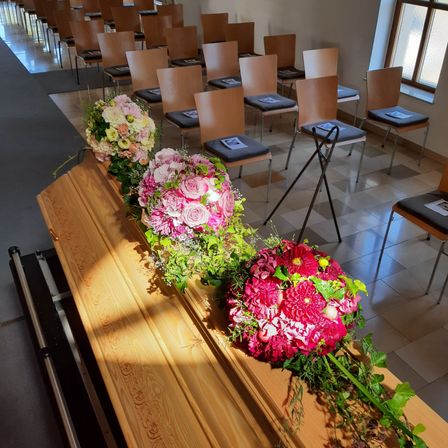 Ein hölzerner Sarg, geschmückt mit rosa und grünen Blumenarrangements, steht in einem Raum mit gefliestem Boden und aufgereihten Stühlen. Jeder Stuhl hat eine schwarze Sitzfläche und ein weißes Papier obenauf.