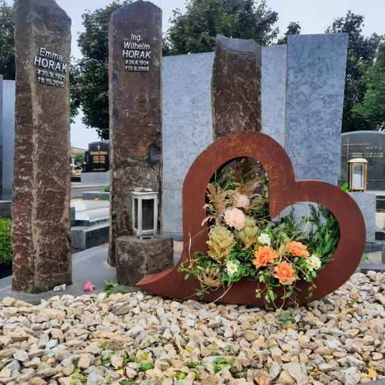 Ein Friedhof mit Grabsteinen für Emma Horak und Ing. Wilhelm Horak. Es gibt eine herzförmige Blumenarrangement mit orangefarbenen und weißen Blumen.