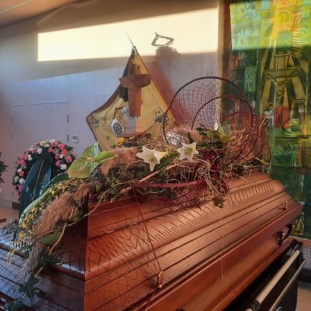 Ein hölzerner Sarg, bedeckt mit weißen und gelben Blumen und grünen Blättern, wird in einem Raum ausgestellt. Über dem Sarg befindet sich eine Flagge mit einem Kreuz. Der Raum hat eine weiße Wand und eine Tür.