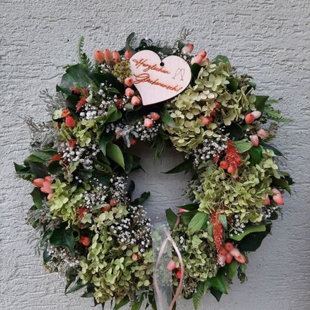 Ein Kranz mit grünen Blättern, roten Beeren und weißen Blumen hängt an einer Wand. Ein Herz mit den Worten Herzlichen Gluckwunsch ist in der Mitte.