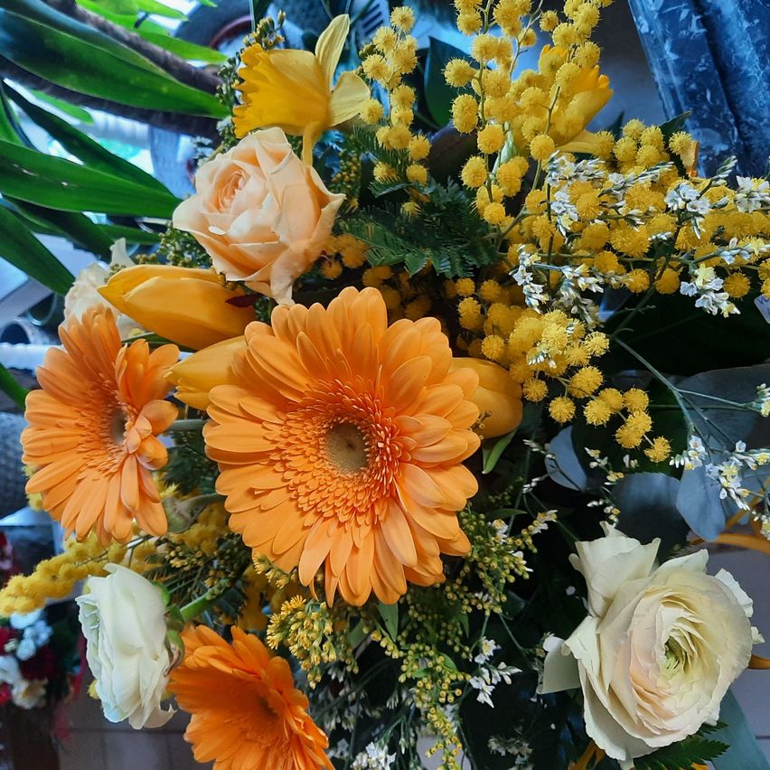 A vibrant bouquet features orange and yellow flowers, including daisies and roses, surrounded by green leaves and a soft blue background.