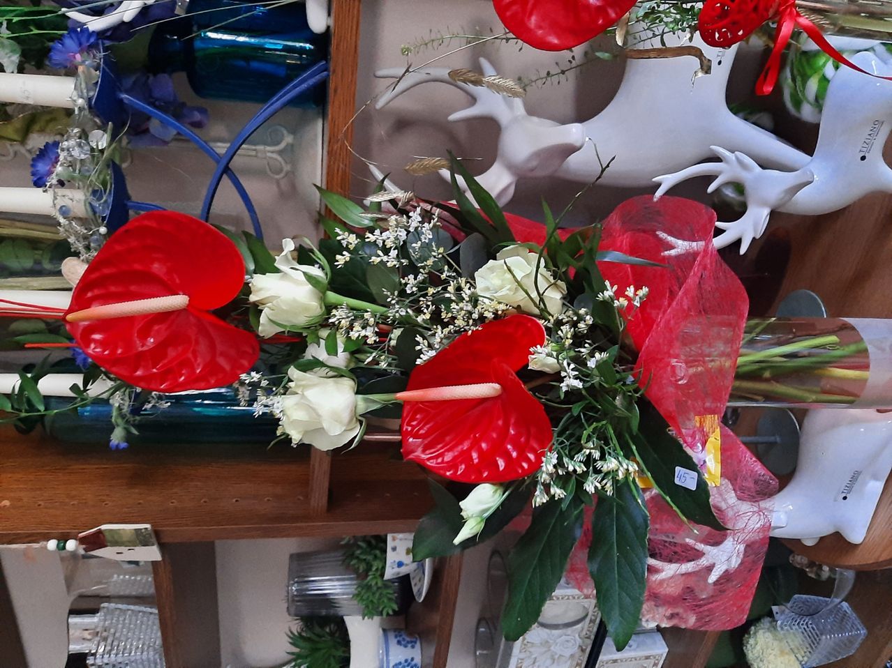 A vibrant floral arrangement features red anthuriums and white roses, surrounded by greenery. Displayed on a wooden shelf, it is wrapped in red plastic and accompanied by other decorative items.