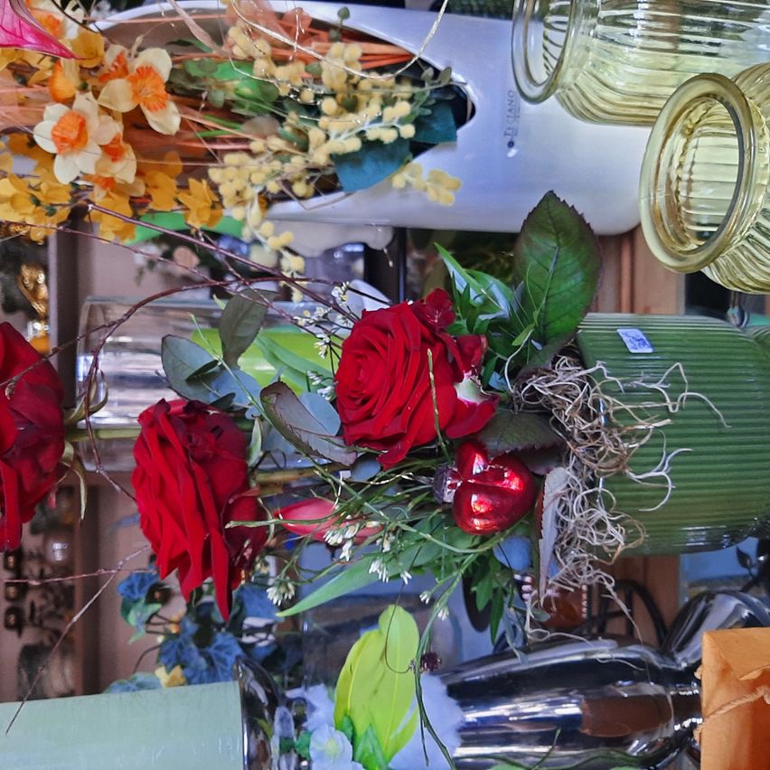 A floral arrangement featuring red roses and yellow daffodils in a green vase. Other decorative items and flowers are in the background.