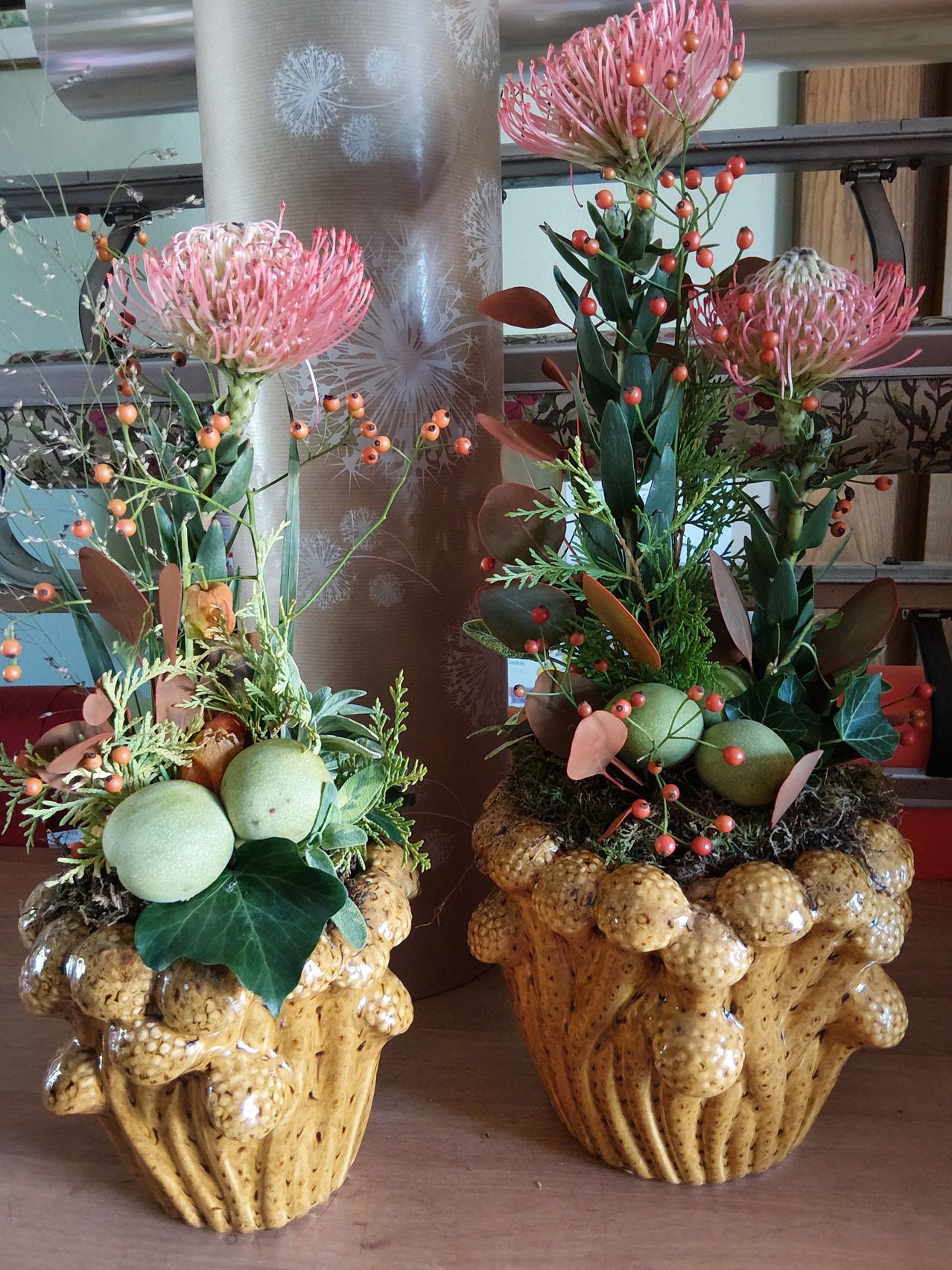 Zwei dekorative Vasen, gefüllt mit einer Mischung aus Blumen, Blättern und Früchten. Die Vasen haben strukturierte Oberflächen und stehen auf einem Holztisch.