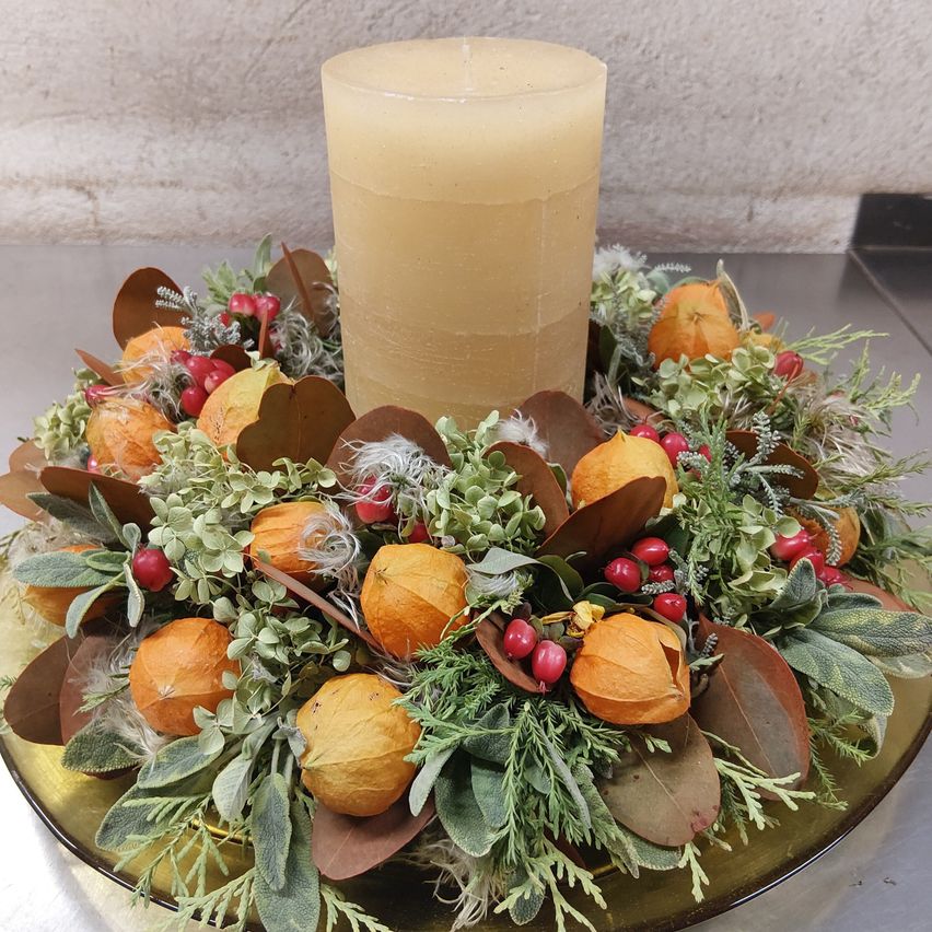 Eine goldene Kerze steht in der Mitte eines Kranzes aus getrockneten Blättern, kleinen orangefarbenen Früchten und roten Beeren auf einem goldenen Teller.