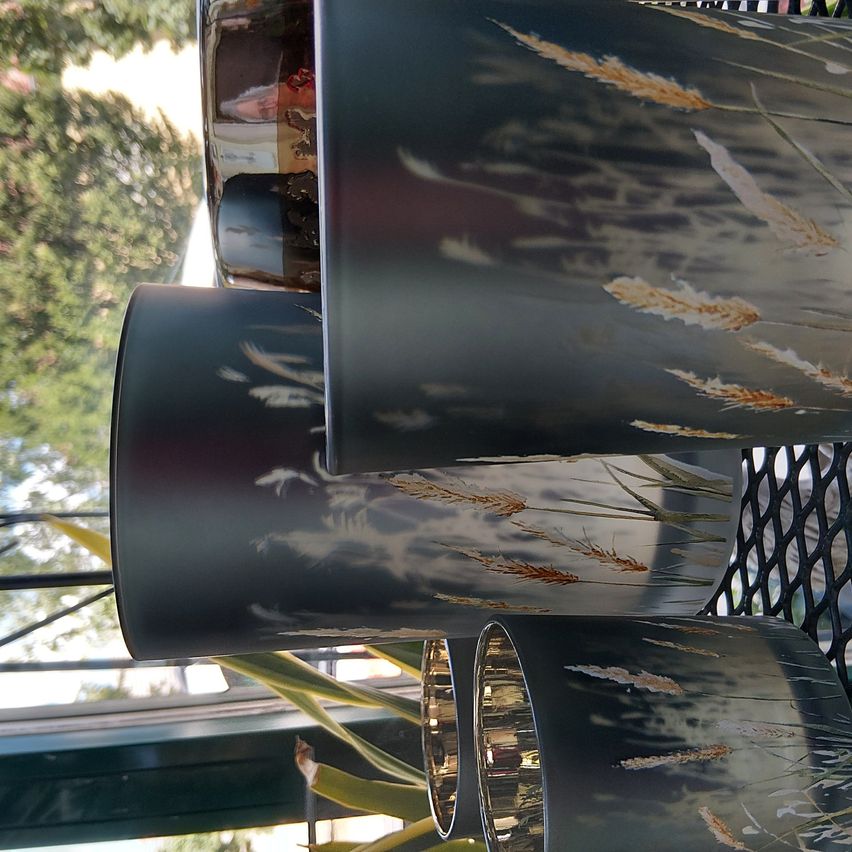 A close-up view of three cylindrical vases with metallic reflections, displaying wheat-like patterns. They rest on a black mesh surface, with blurred greenery and trees in the background.