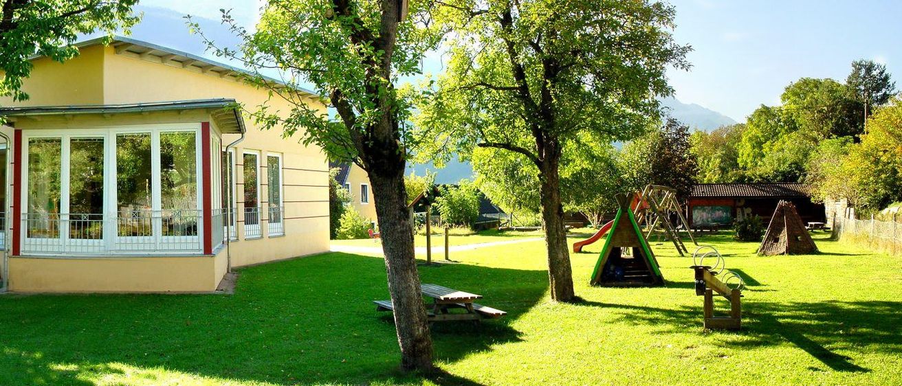 Ein hellgelbes Haus mit einer Veranda aus Glas und Metallgittern ist von grünem Gras und Bäumen umgeben. Ein hölzerner Picknicktisch mit Bänken steht im Garten.