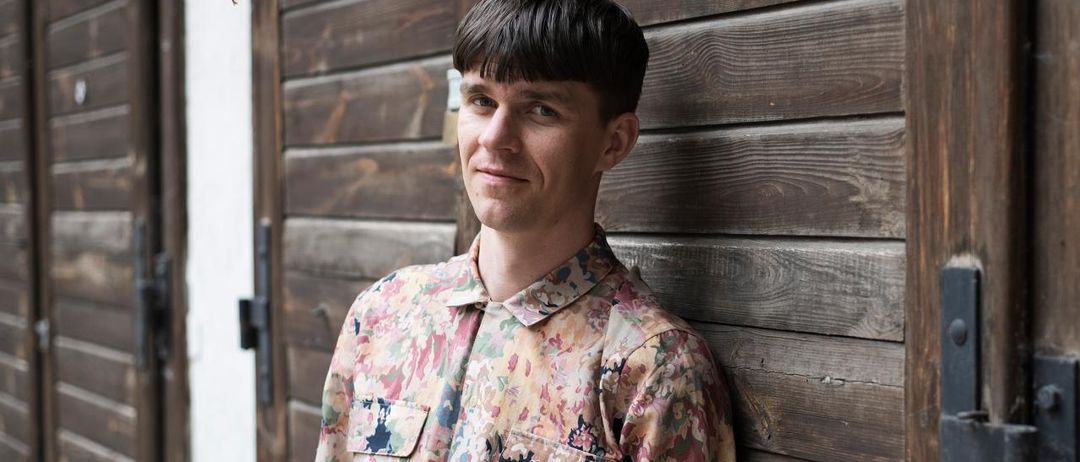 Ein Mann mit kurzen Haaren und einem Blumenhemd steht lächelnd und posiert für ein Foto an einer Holzwand.