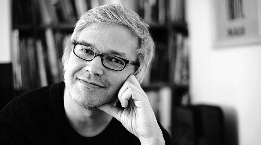 Ein Mann mit einer Brille und einem schwarzen Hemd sitzt in einem Raum und lächelt. Er hat seine rechte Hand am Kinn. Er posiert wahrscheinlich für ein Foto. Dahinter steht ein Bücherregal mit vielen Büchern.