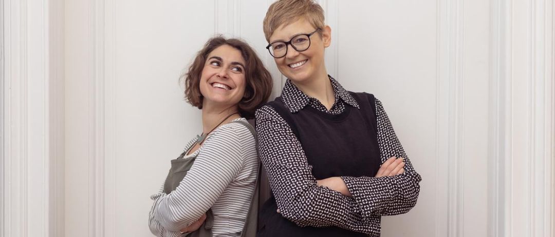 Zwei Frauen stehen lächelnd vor einer weißen Tür. Eine trägt ein gestreiftes Shirt und eine Schürze, die andere ein schwarzes Weste mit Brille.