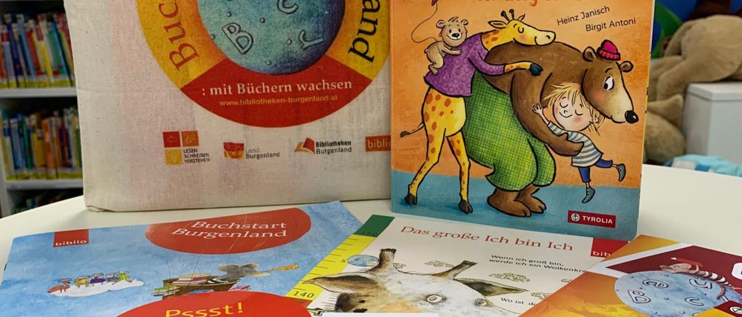 Ein weißer Beutel mit den Worten Buchstart Burgenland darauf. Mehrere Bücher, einschließlich Das mag ich, stehen auf einem Tisch. Die Bücher haben farbenfrohe Illustrationen und sind in einer Reihe angeordnet. Ein Plüschtier liegt auf dem Beutel. Im Hintergrund befinden sich Bücherregale.