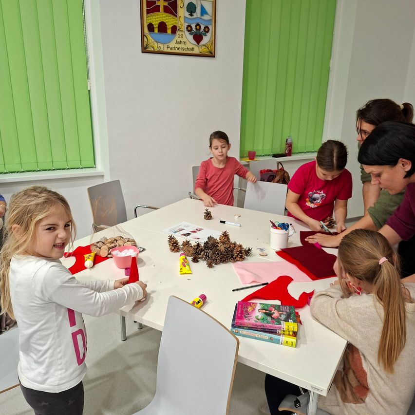 Mehrere Kinder und Erwachsene arbeiten gemeinsam an einem Tisch und basteln mit Materialien wie Tannenzapfen, Papier und Kleber. Der Raum hat grüne Jalousien und ein gerahmtes Bild an der Wand.