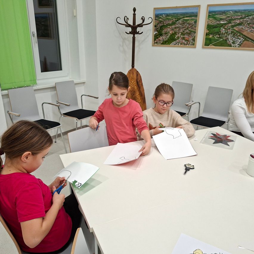 Vier junge Mädchen sitzen um einen Tisch herum und beschäftigen sich mit einer Bastelaktivität. Sie haben ausgeschnittene Formen auf dem Tisch, Scheren und Papiere. Hinter ihnen hängen zwei gerahmte Bilder an der Wand.