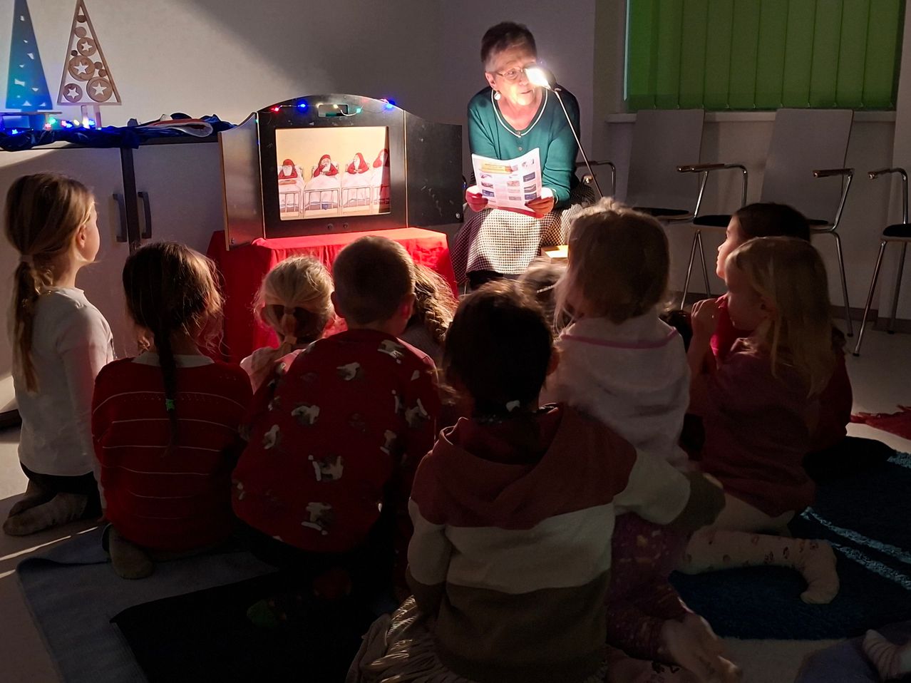 Eine Frau liest ein Buch unter einer Lampe, während Kinder vor einer Projektionsleinwand sitzen und aufmerksam zuschauen.