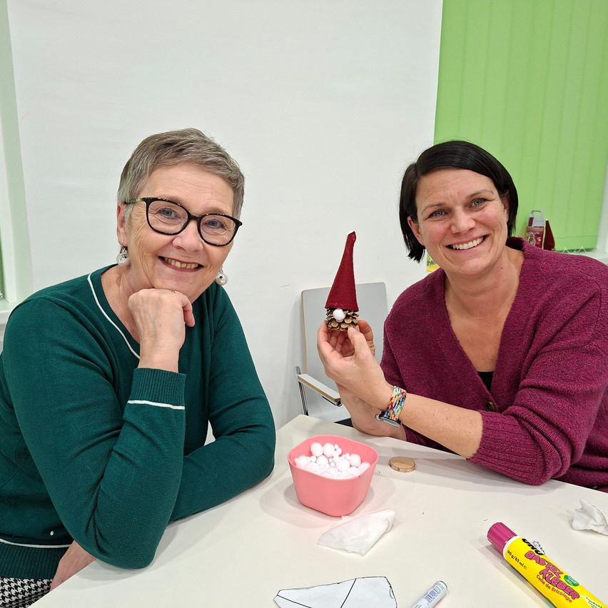 Zwei Frauen lächeln und posieren für ein Foto, während sie um einen Tisch mit Bastelmaterial sitzen. Eine hält ein kleines, rot-konisches Bastelobjekt.