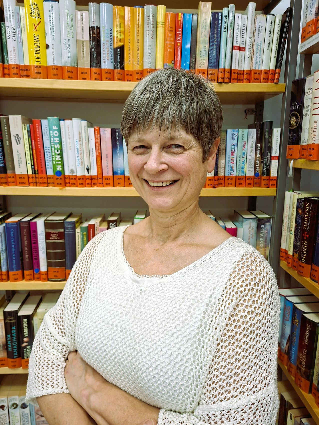 Eine Frau mit kurzen grauen Haaren, lächelt und steht vor einem vollgepackten Bücherregal.