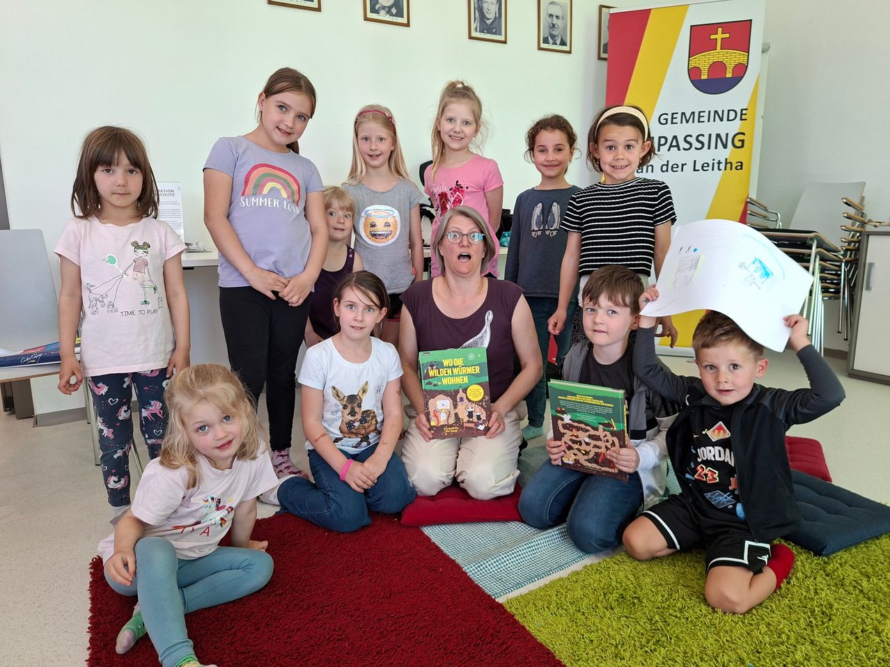 Eine Gruppe von Kindern und Erwachsenen posiert für ein Foto. Die Frau in der Mitte hält ein Buch. Einige Kinder halten Bücher. Die Kinder tragen Freizeitkleidung. Der Raum hat eine weiße Wand mit Bilderrahmen und einem Banner.