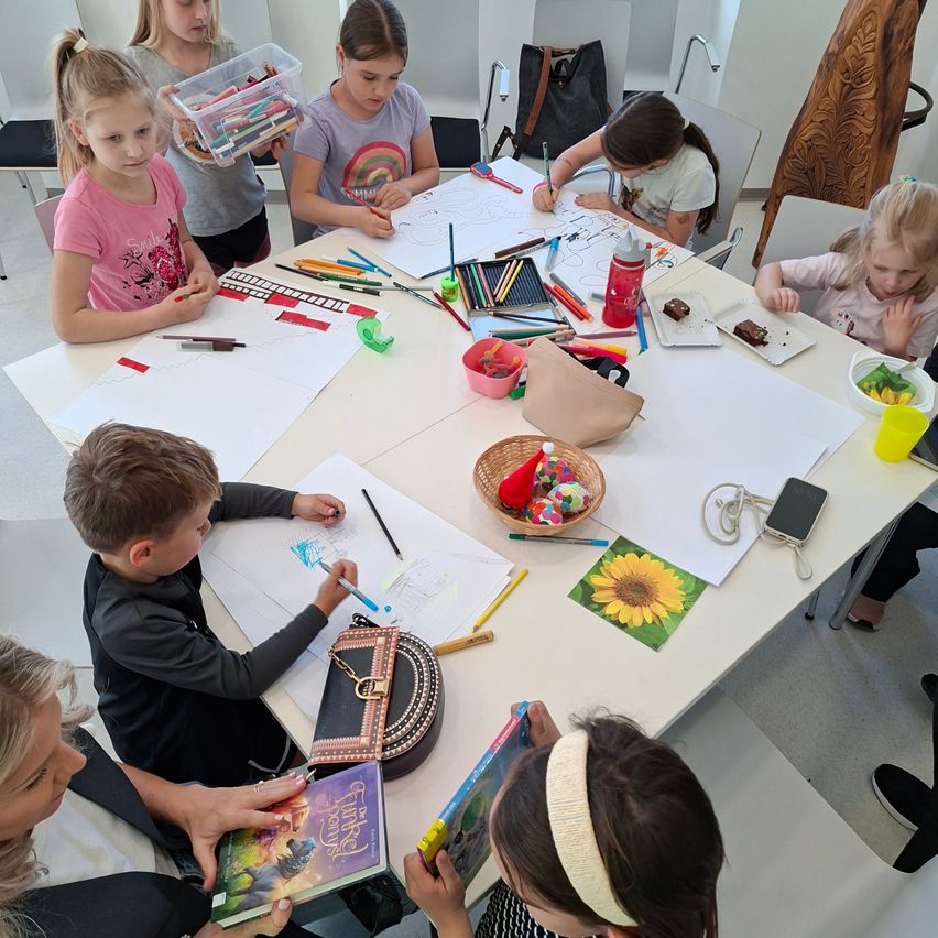 Kinder in einem Klassenzimmer, um einen Tisch herum sitzend, sind mit Zeichenaktivitäten beschäftigt. Eine Vielzahl von Malutensilien und Zeichnungen sind auf dem Tisch. Eine erwachsene Frau ist anwesend, wahrscheinlich eine Lehrerin, die die Aktivität beaufsichtigt.