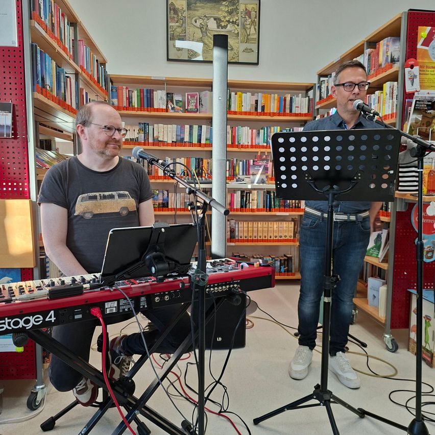 Zwei Männer treten in einer Bibliothek auf. Einer spielt Klavier, während der andere steht und in ein Mikrofon singt.