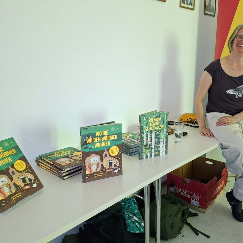 Eine Frau sitzt auf einer Bank mit Büchern auf einem Tisch vor ihr. Die Bücher heißen 'Wo die wilden Würmer wohnen' und 'Fin Baum'.