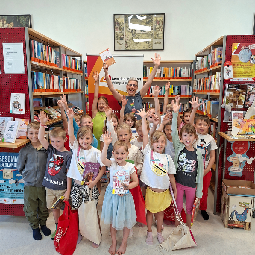 Eine Gruppe junger Kinder in einer Bibliothek mit Büchern, die ihre Hände heben. Sie posieren für ein Foto. Ein Mann im Hintergrund hält ein Buch und lächelt.