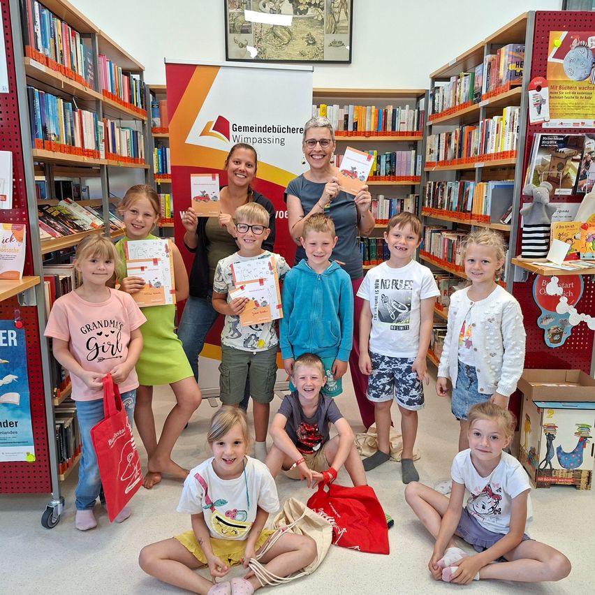 Eine Gruppe von Kindern mit Erwachsenen in einer Bibliothek, die Bücher halten und lächeln. Zwei Frauen stehen hinter ihnen mit Büchern. Regale mit Büchern umgeben sie.