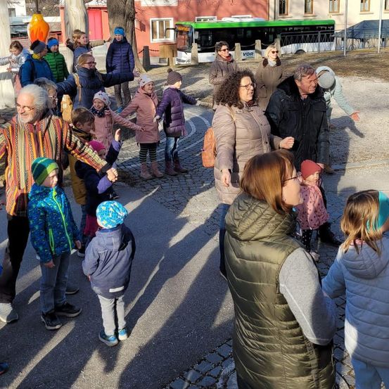 Eine Gruppe von Erwachsenen und Kindern, einige mit Mützen und Brillen, stehen zusammen in einem Außenbereich, möglicherweise einem Park, mit einem Bus und Gebäuden im Hintergrund.