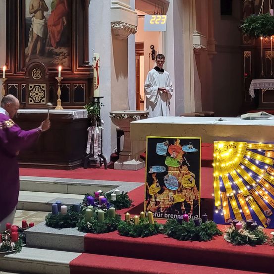 Ein Priester steht in einer Kirche mit einem geschmückten Altar davor. Er hält eine Glocke, und ein anderer Priester steht hinter ihm. Der Altar hat einen Weihnachtsbaum aus Lichtern und ein Bild von Jesus.