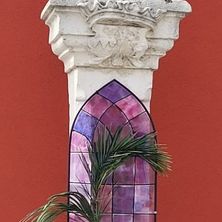 Eine weiße Vogelskulptur auf einem Pfeiler mit einem darunterliegenden Buntglasfenster, das ein Palmblatt und das Wort 'Frohe' zeigt.