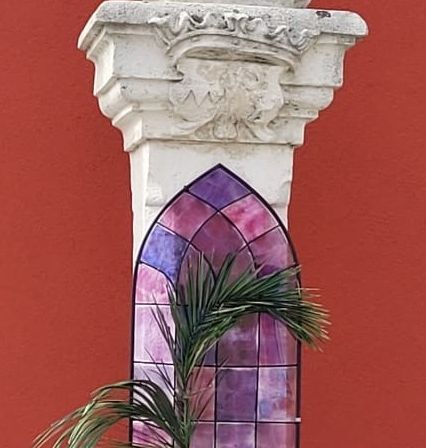 Eine weiße Vogelskulptur auf einem Pfeiler mit einem darunterliegenden Buntglasfenster, das ein Palmblatt und das Wort 'Frohe' zeigt.