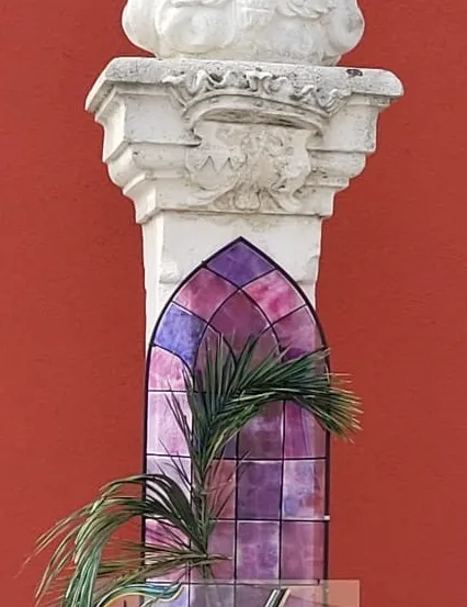 Eine weiße Vogelskulptur auf einem Pfeiler mit einem darunterliegenden Buntglasfenster, das ein Palmblatt und das Wort 'Frohe' zeigt.