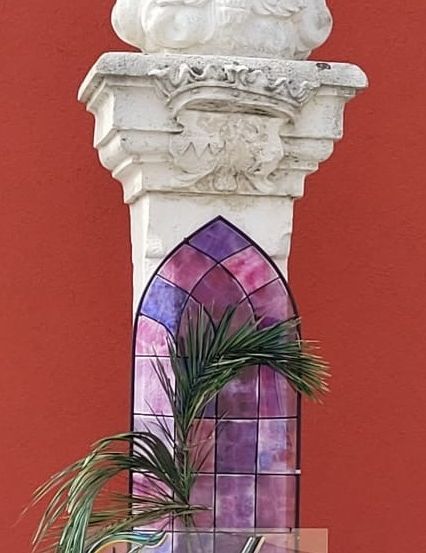 Eine weiße Vogelskulptur auf einem Pfeiler mit einem darunterliegenden Buntglasfenster, das ein Palmblatt und das Wort 'Frohe' zeigt.