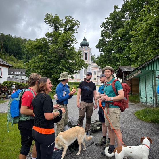 Eine Gruppe von Wanderern mit Rucksäcken steht auf einer Straße vor einer Kirche, mit zwei Hunden an der Leine. Bäume und Gebäude im Hintergrund.