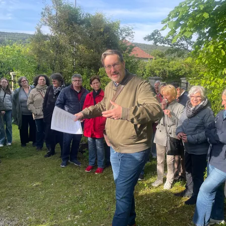 Ein Mann steht im Freien mit einem Papier und spricht eine Gruppe von Menschen in lässiger Kleidung an einem sonnigen Tag an.