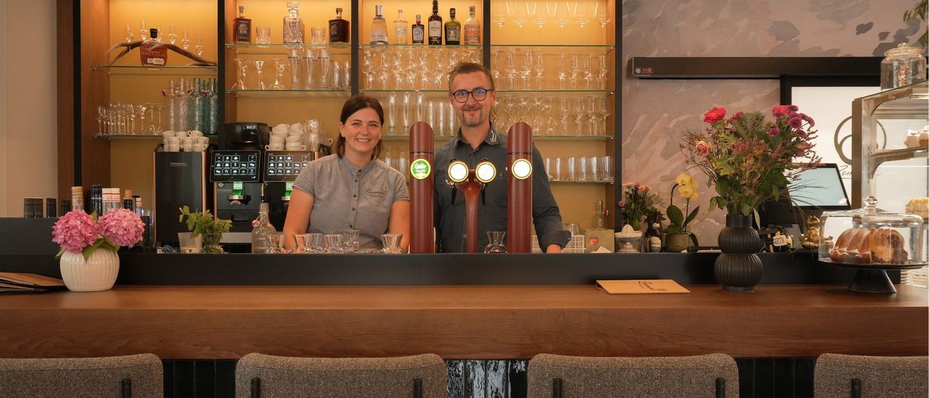 Zwei Personen stehen hinter einer Bar mit Regalen voller Spirituosen und Gläsern. Eine Frau lächelt in die Kamera, während ein Mann wegschaut. Die Bar hat eine Holztheke und zwei Hocker.