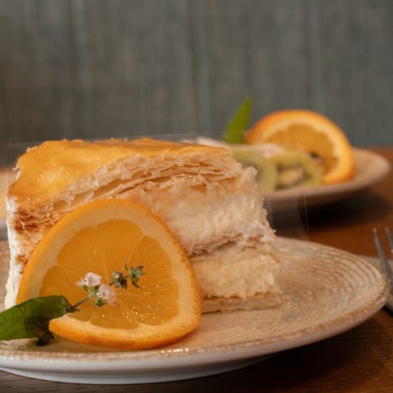 Ein Dessert mit Schichten aus Teig und Sahne, garniert mit einer Orangenscheibe und einer Blume, auf einem Teller mit einer Gabel.