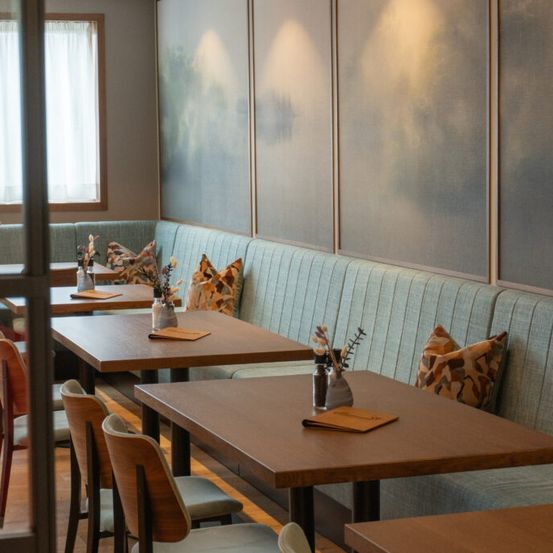 Ein modernes Restaurant mit Holztischen und Stühlen. Die Tische haben Vasen mit Blumen und Menüs. Die Stühle sind um die Tische herum angeordnet. Die Wand hat große Gemälde. Der Raum hat ein Fenster mit weißen Vorhängen.