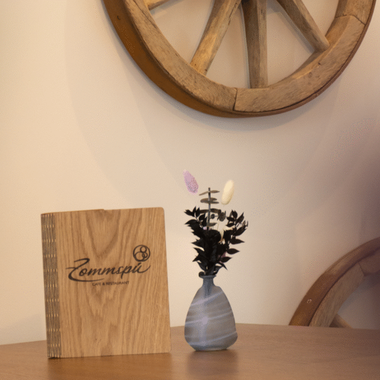 Ein Holzmenübuch und eine Vase mit lila und weißen Blumen stehen auf einem Tisch. Ein hölzerne Rad ist an der Wand darüber montiert.
