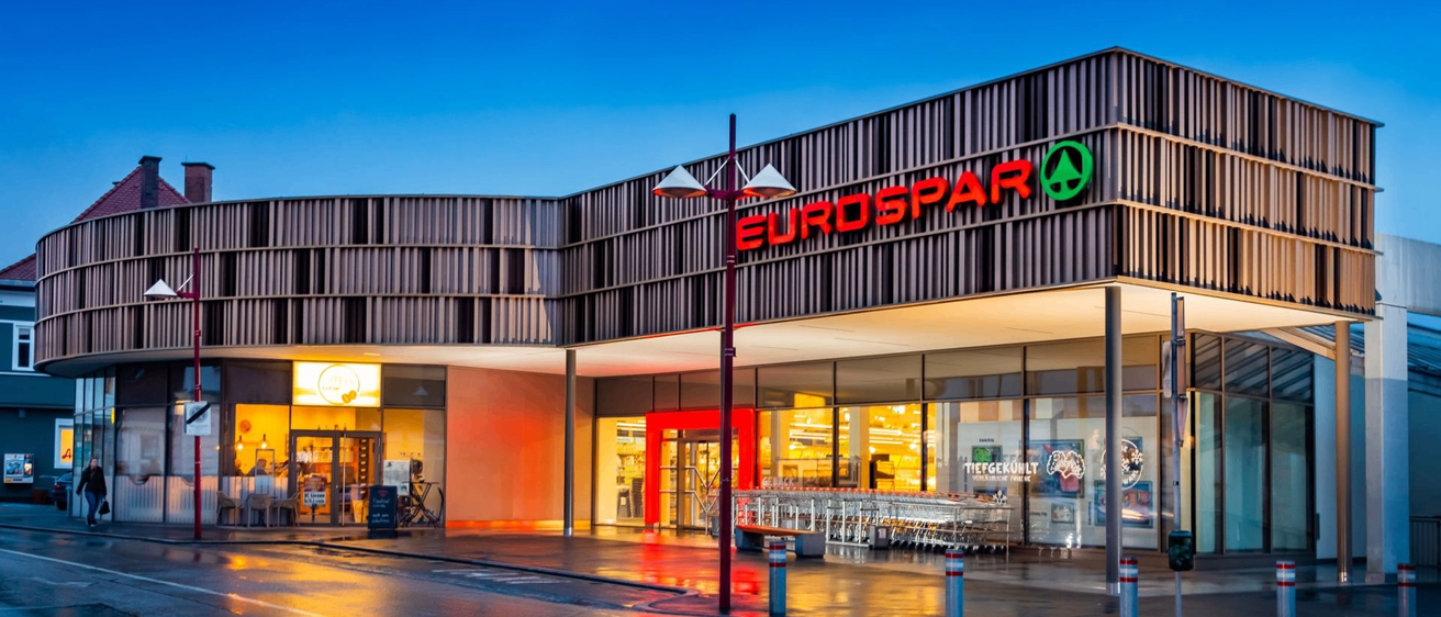 Eurospar Supermarkt mit rotem und grünem Schild, bei Nacht beleuchtet. Helle Lichter reflektieren auf der nassen Straße.