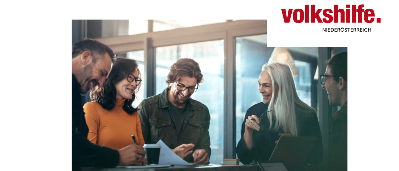 Vier Menschen sitzen lächelnd um einen Tisch herum und diskutieren. Zwei Frauen und zwei Männer, einer hält einen Stift und Papier, der andere einen Laptop.