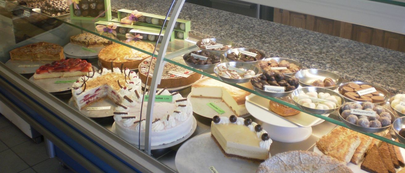 Eine Glasvitrine mit einer Auswahl an Kuchen und Desserts, darunter mehrschichtige Kuchen, Käsekuchen und verschiedene Gebäcke, mit Schokoladen und anderen Süßigkeiten auf dem obersten Regal.
