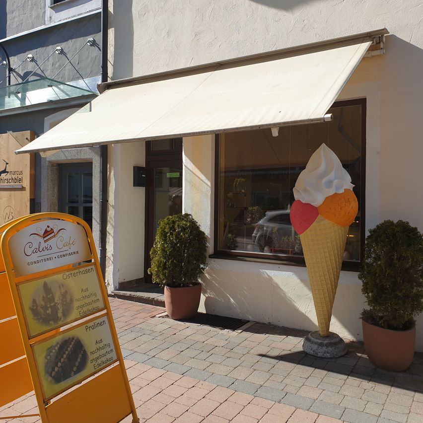 Ein Café-Eingang mit einer großen Eiscreme-Statue davor. Das Café-Schild steht für 'Calis Cafe'. Bepflanzte Töpfe flankieren den Eingang, und ein Auto spiegelt sich im Fenster wider.