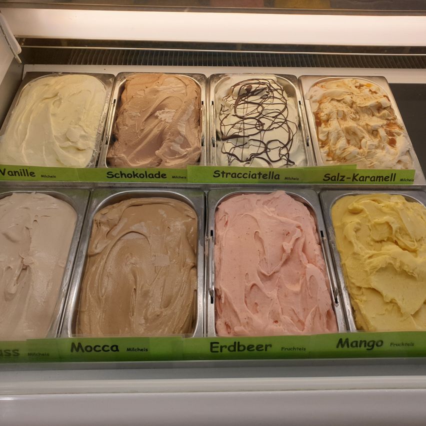 Verschiedene Eissorten sind in einem Gefrierschrank ausgestellt. Die Geschmacksrichtungen umfassen Vanille, Schokolade, Tiramisu, Salz-Karamell, Haselnuss, Erdbeere und Mango.