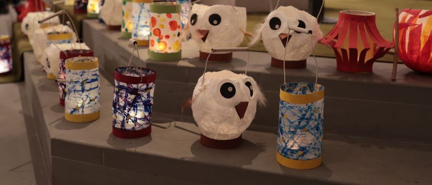 Im Inneren einer Kirche sind auf den Stufen eine Reihe handgefertigter Papierlaternen zu sehen, jede in Form einer Eule. Die Laternen haben verschiedene Farben und Größen.
