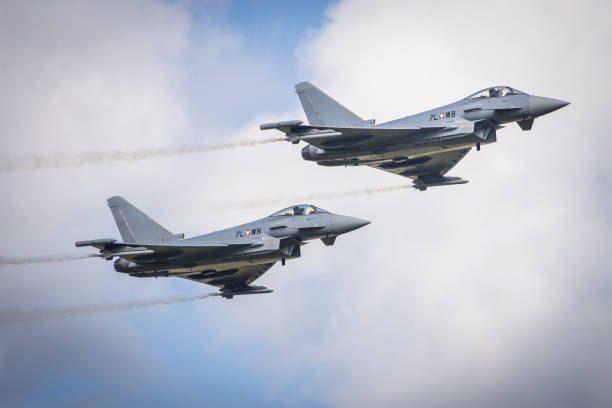 Zwei graue Militärjets fliegen in Formation gegen einen blauen Himmel mit Wolken und hinterlassen Rauchschwaden. Ein Jet hat '7L' an seiner Seite.