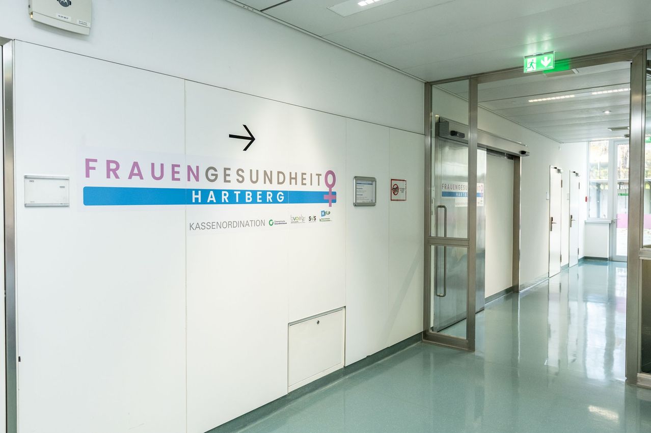 Ein Flur mit weißen Wänden und blauem Boden führt zu einer Tür mit der Aufschrift 'Frauengesundheit Hartberg' und einem Pfeil nach links. Die Wand weist auch ein Schild für Kassenordination auf.