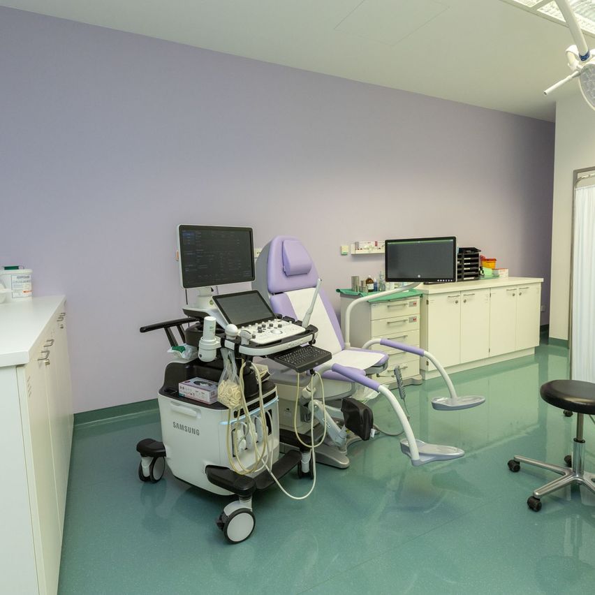 Ein Arztzimmer mit einem Samsung-Ultraschallgerät, einem Stuhl und Monitoren. Schränke, ein Hocker und eine Leuchte sind ebenfalls vorhanden. Der Raum hat einen grünen Boden und lila Wände.