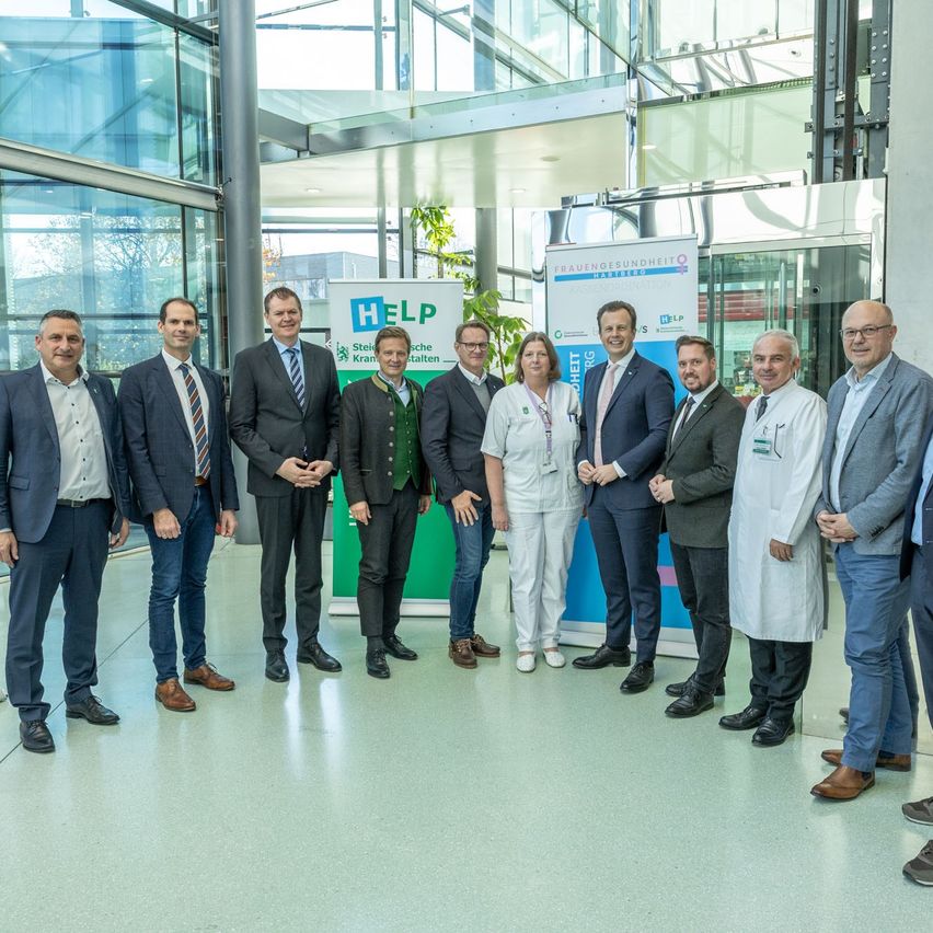 Eine Gruppe von Menschen in formeller Kleidung steht vor einem Banner mit der Aufschrift 'HILFE'. Sie sind bei einer Veranstaltung und posieren für ein Foto.