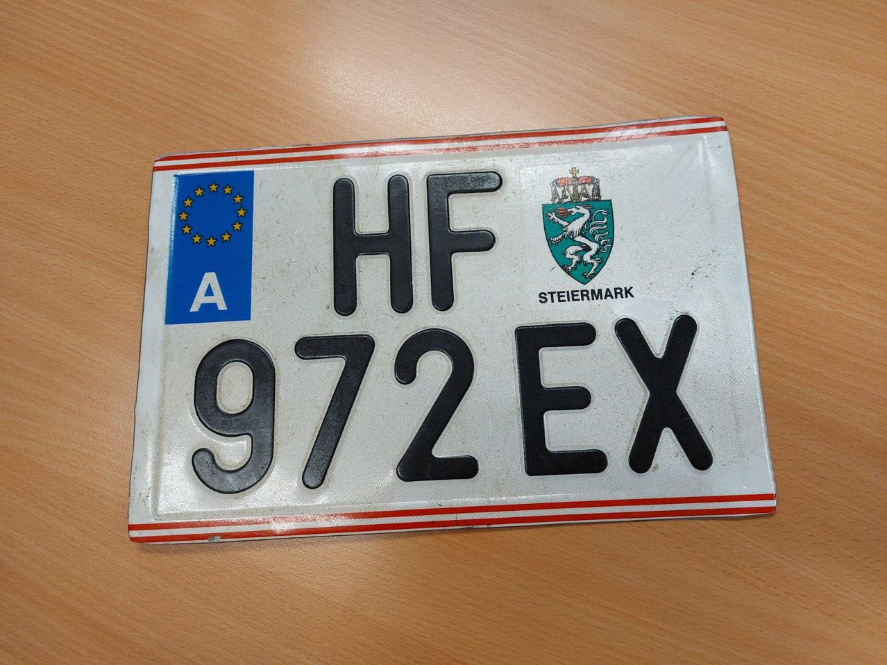 Ein weißer Nummernschild mit schwarzem Text zeigt HF 972EX. Es trägt die EU-Flagge und ein Wappen mit der Aufschrift 'Steiermark'.